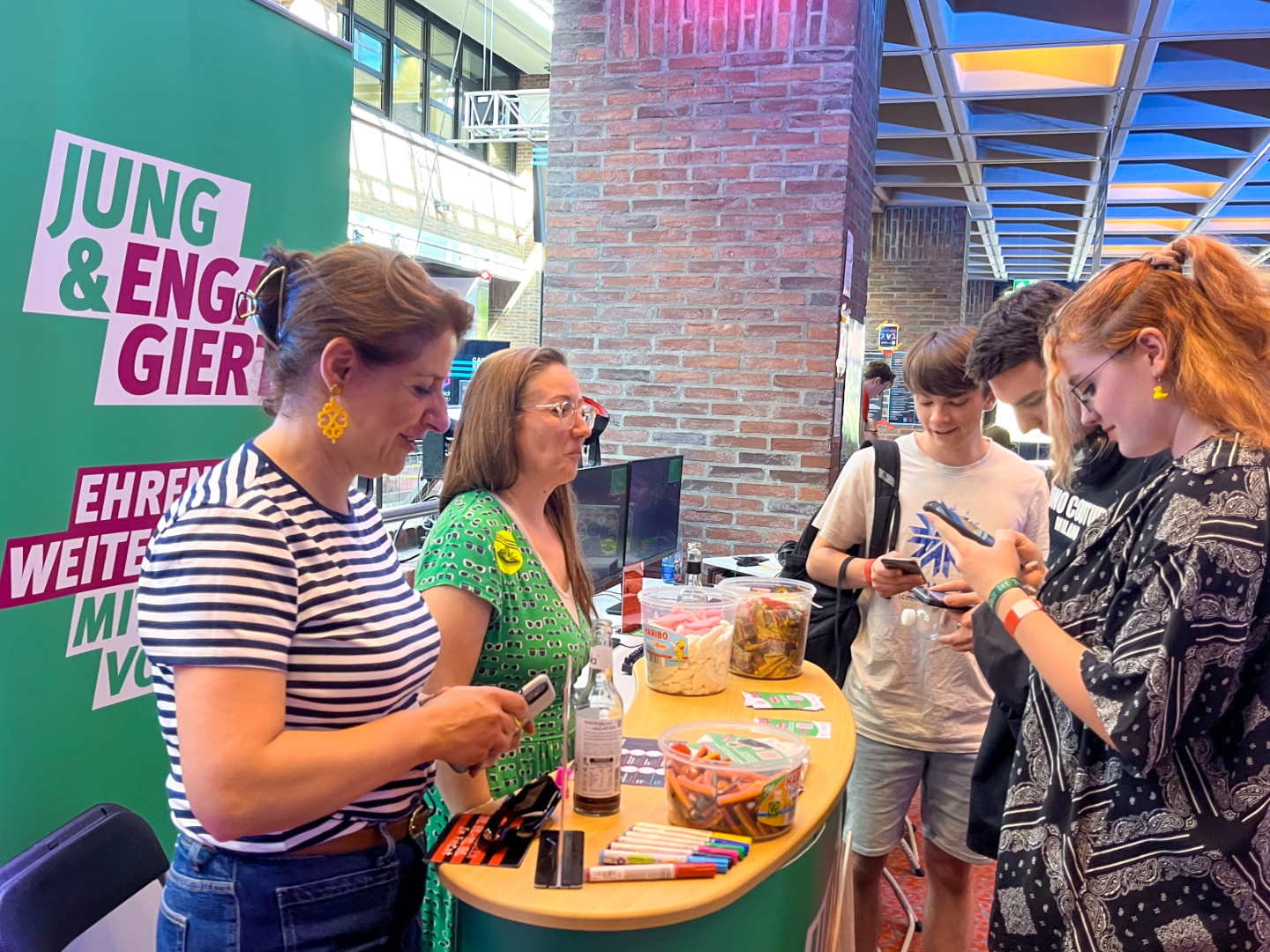Auf einer Veranstaltung von Jung & Engagiert stehen 2 Erwachsene an einem Stand mit Süßigkeiten, Stiften und Flyern. Vor dem Stand stehen 3 junge Menschen, die auf ihre Handys schauen.